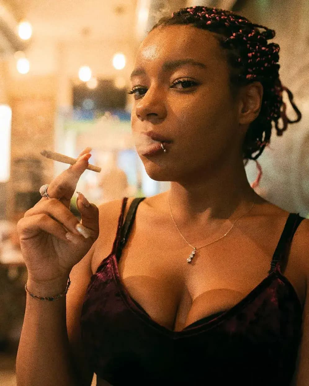 Woman wearing jewelry smoking a cigar in a relaxed lounge setting with warm lighting.