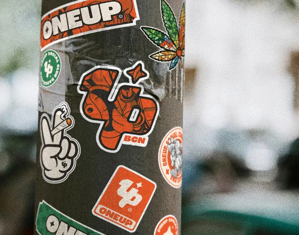 Sticker-covered street pole featuring ONEUP logo decals and urban brand artwork in Barcelona.